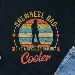 Könnte beinhalten: Schwarzes T-Shirt mit einer Grafik im Vintage-Stil einer Person, die ein einrädriges Elektro-Skateboard fährt. Die Grafik lautet "Onewheel Dad Like a Regular Dad But Cooler".
