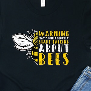 Può includere: Maglietta nera con un'immagine di un'ape gialla e nera e il testo "WARNING MAY SPONTANEOUSLY START TALKING ABOUT BEES".