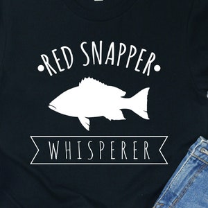 May include: Black t-shirt with white text that reads "Red Snapper Whisperer" and a white silhouette of a fish.