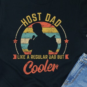 Puede incluir: Camiseta negra con un gráfico de estilo vintage que dice "Host Dad Like a Regular Dad But Cooler". El gráfico presenta una silueta de una persona sosteniendo un micrófono frente a una puesta de sol.
