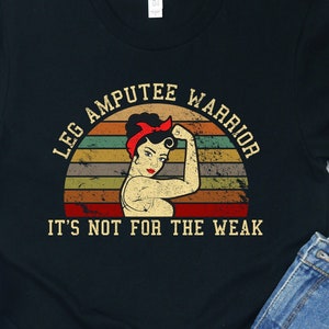May include: Black t-shirt with the text "LEG AMPUTEE WARRIOR" above a vintage-style illustration of a woman flexing her bicep. The text "IT'S NOT FOR THE WEAK" is below the illustration. White sneakers and denim shorts are also in the image.