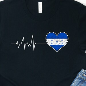 Peut inclure: T-shirt noir avec un graphique de battement de cœur blanc et un cœur bleu avec le drapeau du Honduras à l'intérieur. Le cœur est situé sur le côté gauche du t-shirt.