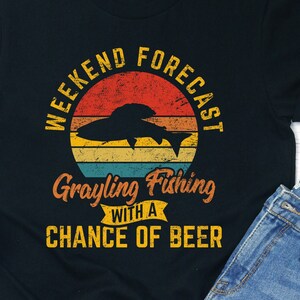May include: Black t-shirt featuring a vintage-style graphic that reads "Weekend Forecast Grayling Fishing with a Chance of Beer". The graphic includes a fish silhouette and a sunset with a retro colour scheme.