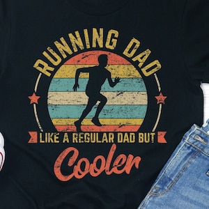 Pode incluir: Camiseta preta com um gráfico de estilo vintage que diz "Running Dad Like a Regular Dad But Cooler". O gráfico apresenta uma silhueta de um corredor sobre um fundo de cor de pôr do sol.