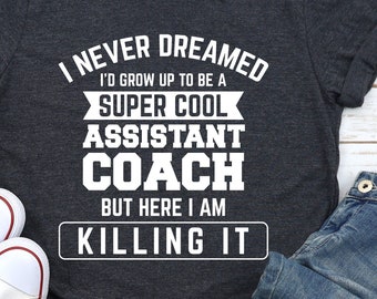 May include: A dark grey t-shirt with white text that reads "I never dreamed I'd grow up to be a super cool assistant coach but here I am killing it". The shirt is lying on a white wooden surface with a pair of white trainers and blue jeans.
