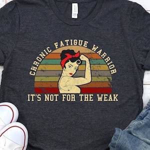 Op de afbeelding: Een donkergrijs T-shirt met een vintage-stijl afbeelding van een vrouw met haar vuist omhoog. De afbeelding zegt "Chronic Fatigue Warrior" en "It's not for the weak".
