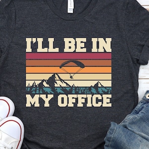 May include: A dark grey t-shirt with a vintage-style graphic of a paraglider flying over mountains. The text "I'll be in my office" is printed on the shirt.
