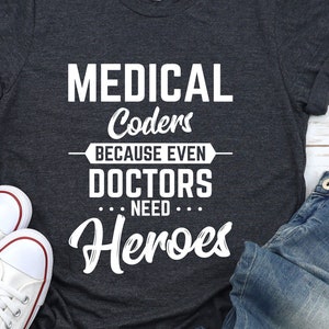 May include: A dark grey t-shirt with white text that reads "MEDICAL Coders BECAUSE EVEN DOCTORS NEED Heroes". The shirt is laid out on a light wood surface with a pair of white canvas shoes and blue jeans.