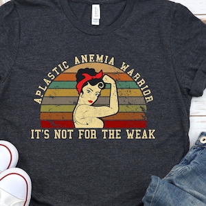Può includere: Una maglietta grigia melange scuro con una grafica in stile vintage di una donna con il pugno alzato. La grafica include il testo "Aplastic Anemia Warrior" e "It's not for the weak".
