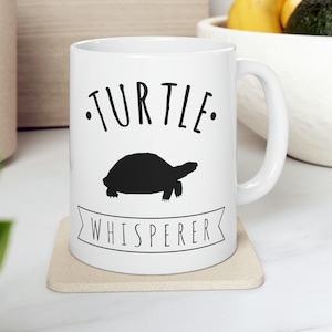 May include: White ceramic coffee mug with a black silhouette of a turtle and the text "TURTLE WHISPERER" printed on it.