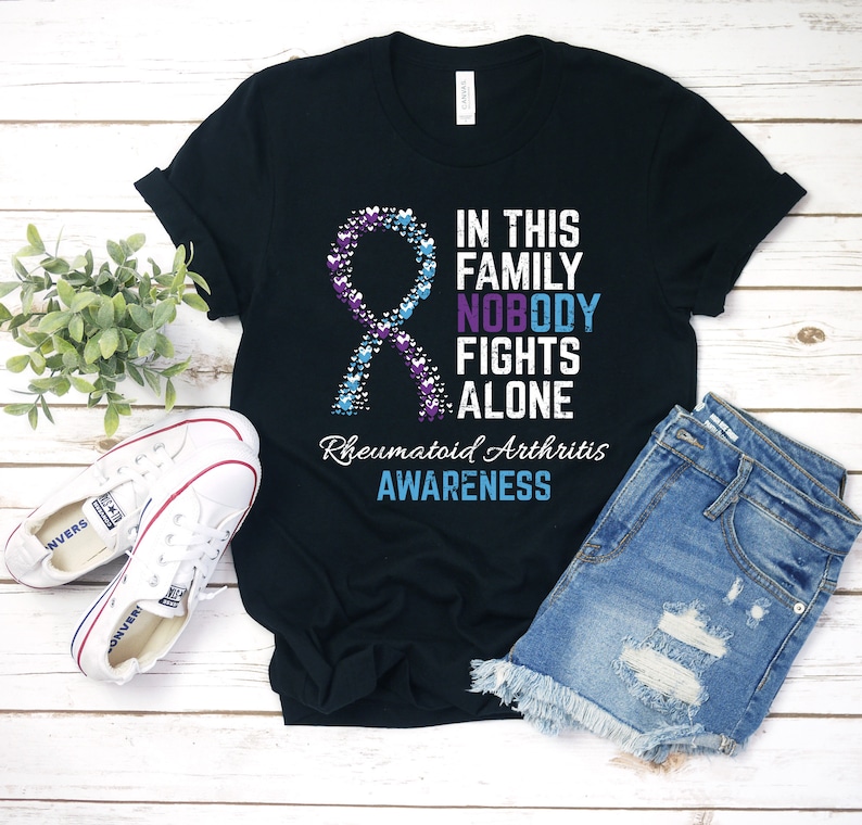 May include: Black t-shirt with a purple and blue ribbon made of hearts. The text "In this family nobody fights alone" and "Rheumatoid Arthritis Awareness" is printed on the shirt.
