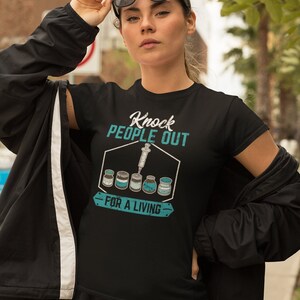 May include: Black t-shirt with a teal graphic that says "Knock People Out - For A Living" with a syringe and vials of medicine.