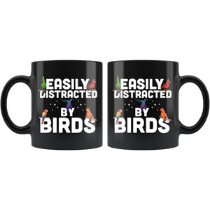 Puede incluir: Dos tazas de cer&aacute;mica negra con el texto "EASILY DISTRACTED BY BIRDS" en blanco. Las tazas presentan ilustraciones coloridas de varios p&aacute;jaros. Las tazas est&aacute;n dise&ntilde;adas para los amantes de las aves y son perfectas para caf&eacute; o t&eacute;.