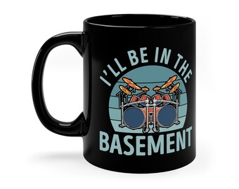 Op de afbeelding: Zwarte koffiemok met een witte afbeelding van een drumstel en de tekst "I'll be in the basement".