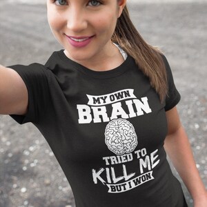 Puede incluir: Camiseta negra con texto blanco que dice "My Own Brain Tried To Kill Me But I Won". El texto est&aacute; rodeado de una pancarta y un gr&aacute;fico de cerebro est&aacute; en el centro.