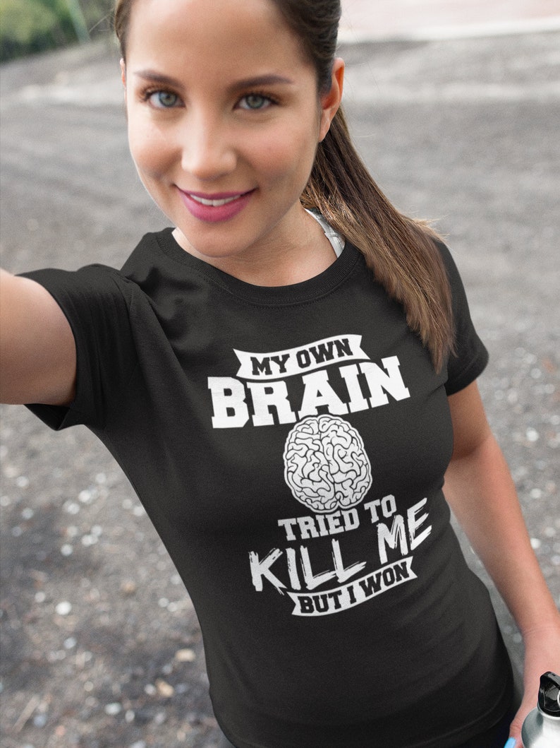Puede incluir: Camiseta negra con texto blanco que dice "My Own Brain Tried To Kill Me But I Won". El texto est&aacute; rodeado de una pancarta y un gr&aacute;fico de cerebro est&aacute; en el centro.
