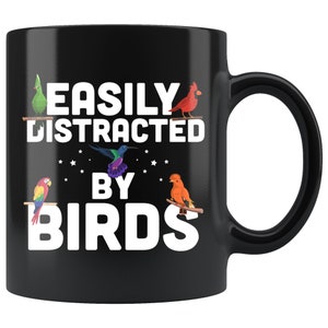 Puede incluir: Taza de cer&aacute;mica negra con el texto "EASILY DISTRACTED BY BIRDS" en blanco. La taza presenta ilustraciones coloridas de varias aves, incluyendo un loro, un cardenal y un colibr&iacute;, posadas en ramas.