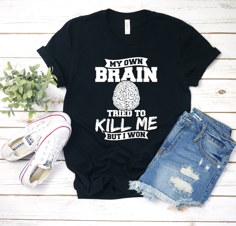 Puede incluir: Camiseta negra con texto blanco que dice "My Own Brain Tried To Kill Me But I Won". El texto est&aacute; rodeado de un contorno blanco. Un contorno blanco de un cerebro est&aacute; por encima del texto.