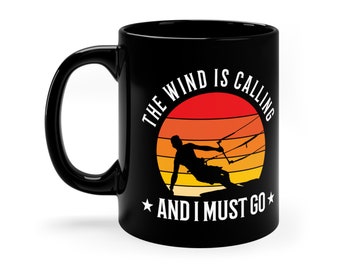 May include: Black coffee mug with a white graphic of a silhouette of a person kitesurfing in front of a sunset with the text "The wind is calling and I must go."