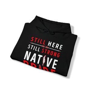 May include: Black hooded sweatshirt with the text "Still Here Still Strong Native Pride" in white with red accents. The text is surrounded by two arrows pointing to the right.