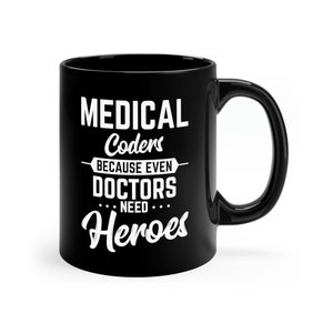 Op de afbeelding: Zwarte keramische koffiemok met witte tekst die "MEDICAL Coders BECAUSE EVEN DOCTORS NEED Heroes" luidt.