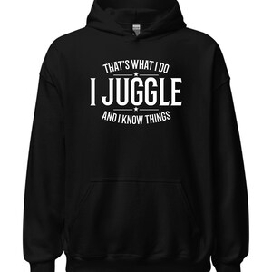 May include: Black hooded sweatshirt with the text "THAT'S WHAT I DO I JUGGLE AND I KNOW THINGS" in white. The sweatshirt has a front pocket and a drawstring hood. A casual, comfortable garment.