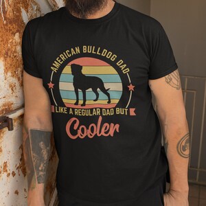 Puede incluir: Una camiseta negra con un gr&aacute;fico de estilo vintage de una silueta de American Bulldog frente a una puesta de sol. El texto dice "American Bulldog Dad - Like a regular dad but cooler".