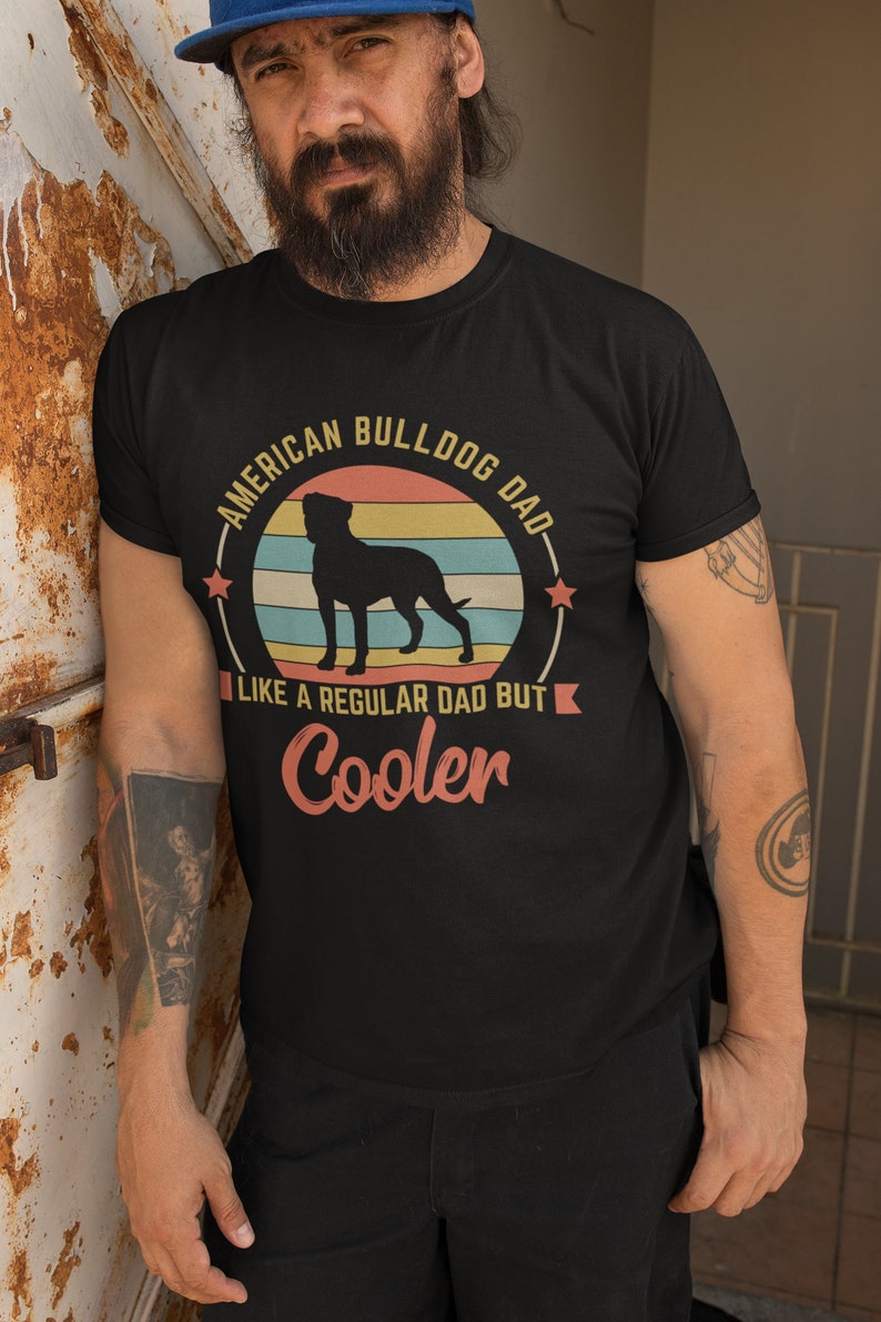 Puede incluir: Una camiseta negra con un gr&aacute;fico de estilo vintage de una silueta de American Bulldog frente a una puesta de sol. El texto dice "American Bulldog Dad - Like a regular dad but cooler".