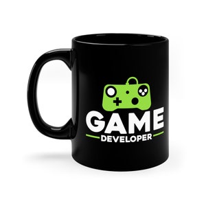 Könnte beinhalten: Schwarze Kaffeetasse mit einem grünen Videospiel-Controller-Motiv und dem Text "GAME DEVELOPER" in Weiß.