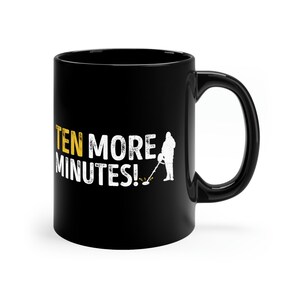 Op de afbeelding: Zwarte keramische mok met een wit silhouet van een persoon die een metaaldetector vasthoudt en de tekst "TEN MORE MINUTES!" in geel.