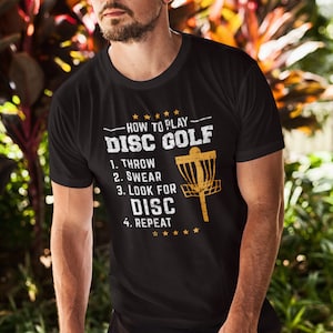May include: A black t-shirt with a gold graphic that says "How to Play Disc Golf" with a list of steps: 1. Throw, 2. Swear, 3. Look for Disc, 4. Repeat. The graphic also includes a disc golf basket.