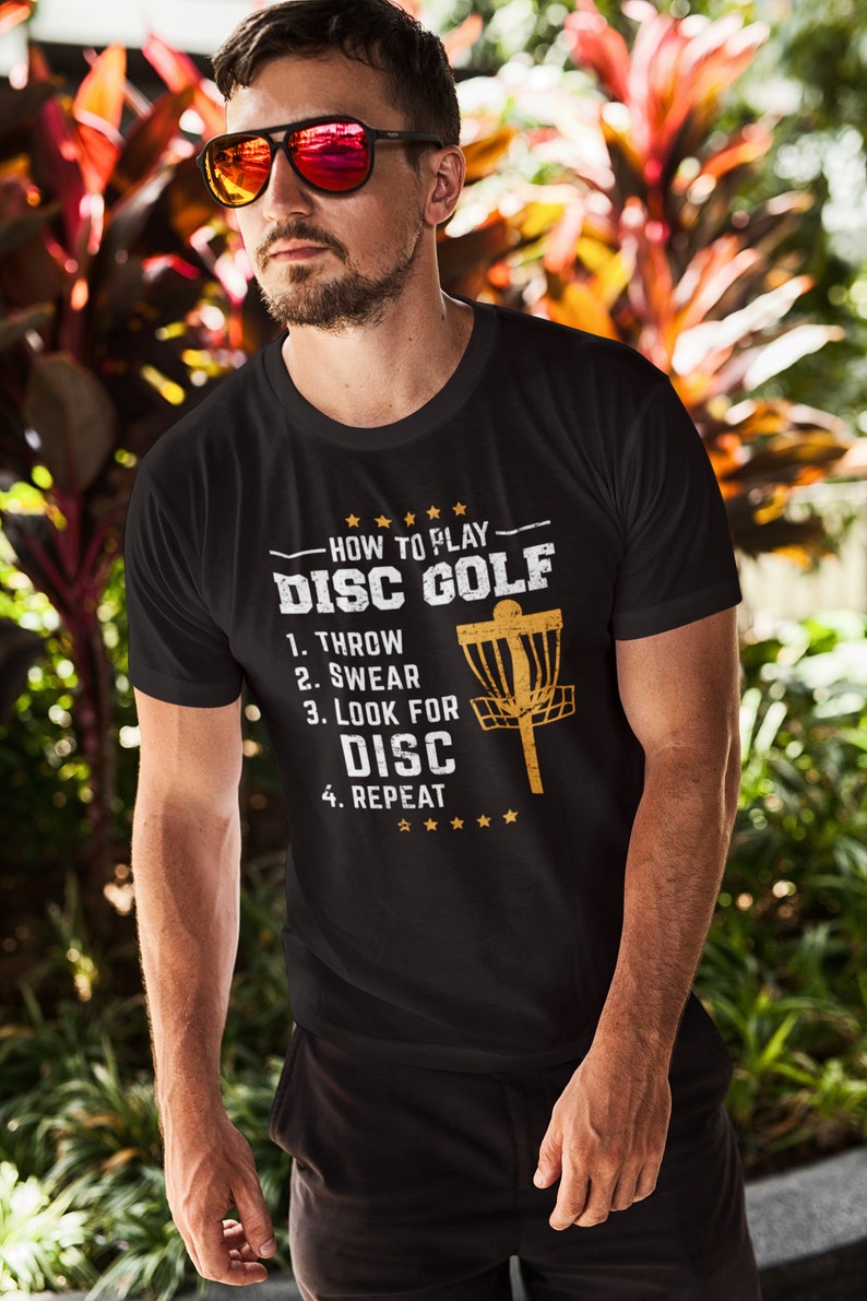 May include: A black t-shirt with a gold graphic that says "How to Play Disc Golf" with a list of steps: 1. Throw, 2. Swear, 3. Look for Disc, 4. Repeat. The graphic also includes a disc golf basket.
