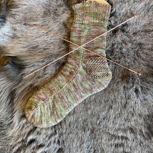 Puede incluir: Un calcetín a rayas verde, rosa y marrón con un talón texturizado. El calcetín está colgado de un gancho con agujas de tejer cruzadas detrás.