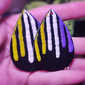May include: A pair of teardrop-shaped earrings with a black base. Each earring features vertical stripes of white, yellow, and purple paint that drip down. The earrings are held in a hand.
