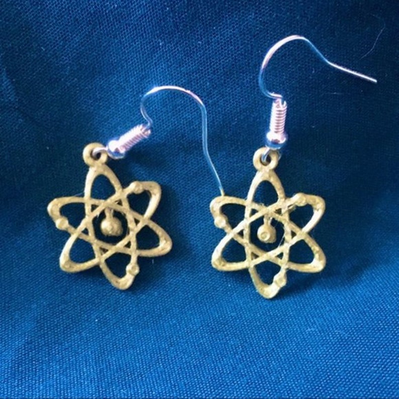 May include: A pair of gold atom earrings with silver hooks. The atom symbol is a stylized representation of an atom with a nucleus and orbiting electrons.