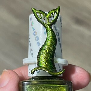 Può includere: Un flacone di smalto per unghie verde con un tappo a forma di coda di sirena olografico. La bottiglia è etichettata "HOLOGRAPHIC".