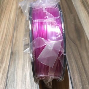 May include: A spool of bright pink filament for 3D printing, wrapped in clear plastic packaging.