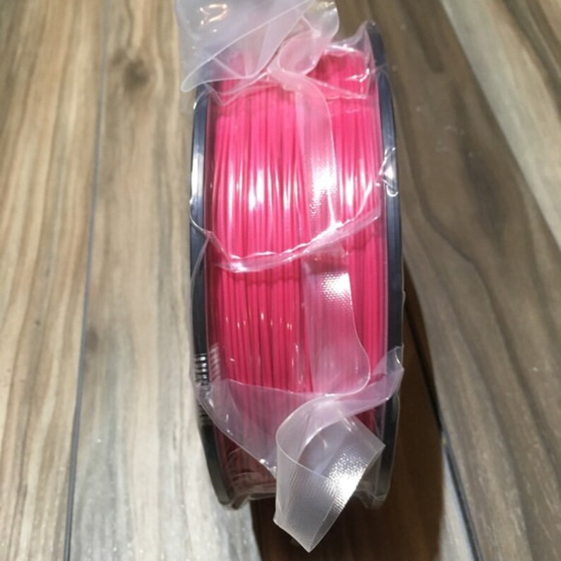May include: A spool of bright pink filament for 3D printing, wrapped in clear plastic and secured with a ribbon.