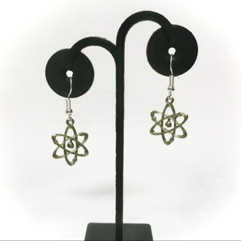 May include: A pair of silver dangle earrings with a green atom charm. The earrings are displayed on a black earring stand.