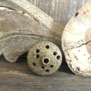 May include: A small, round, green ceramic incense burner with multiple holes for incense sticks. The burner has a speckled glaze and is sitting on a wooden surface.
