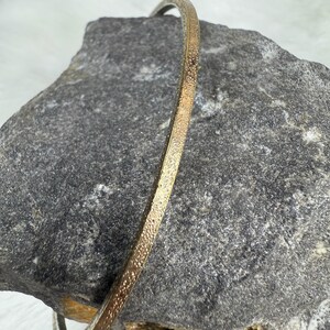 May include: A gold-colored, textured bangle bracelet. The bracelet has a narrow band with a dotted pattern. The jewelry is displayed on a gray rock surface.