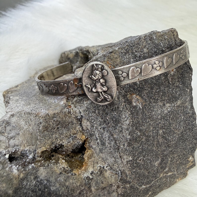 May include: Silver-toned cuff bracelet featuring a Minnie Mouse oval charm. The bracelet has a decorative band with heart and floral patterns. The bracelet is displayed on a gray rock.