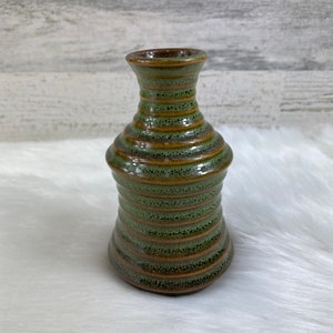 May include: A small, green and brown ceramic vase with a textured, striped design. The vase has a narrow neck and a wide base.