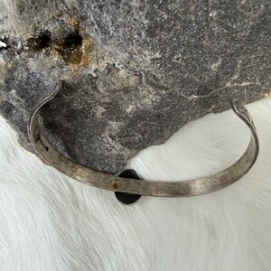 May include: A silver-toned cuff bracelet with a dark, circular stone set in the center. The bracelet has a slightly curved design and is resting on a gray rock surface. The cuff is likely made of metal and has a vintage or antique appearance.