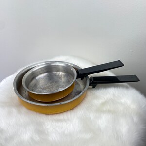 May include: A set of two yellow aluminum frying pans with black plastic handles. The smaller pan is nested inside the larger pan.