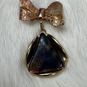 May include: A gold-toned metal brooch featuring a bow at the top with an etched floral pattern. Suspended from the bow is a triangular pendant with a dark, iridescent, and glittery surface encased in a smooth, polished gold-toned metal frame.