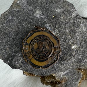 May include: A vintage bronze-toned medal with the text "Black Hawk Archers Corning New York" encircling a central emblem of an eagle. The medal has a decorative border and is set against a textured stone background.
