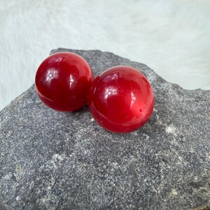 May include: Two glossy, round, red objects rest on a gray, textured surface. The spheres have a smooth, reflective finish, catching the light. The background is a soft, white fabric. The objects appear to be decorative or possibly part of a larger design.
