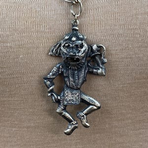 May include: A silver-toned pendant featuring a detailed, stylized figure of a dancing jester. The jester is wearing a hat and holding a staff.
