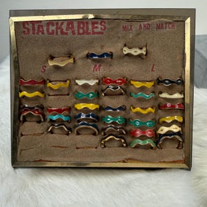 May include: A collection of colorful stackable rings in a display case. The rings are in various colors, including red, yellow, blue, green, and white. The case has the word "STACKABLES" printed on it.